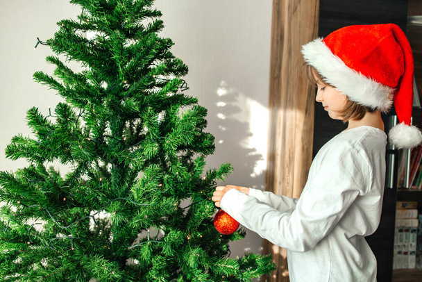 Dziewczyna w sylwestrowym kapeluszu Świętego Mikołaja ubiera choinkę, wiesza zabawki na gałęziach. Przygotowanie do wakacji. - Zdjęcie, obraz