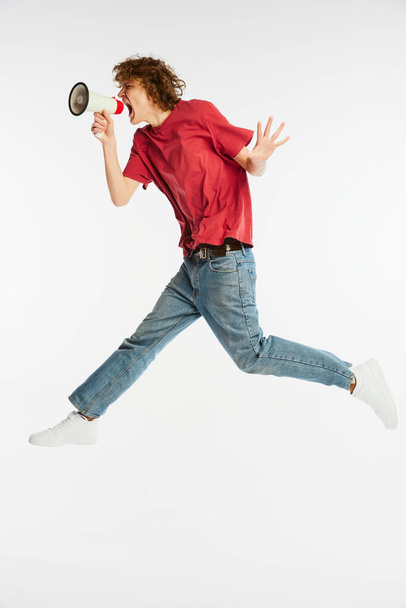 News. Full length portrait of excited young man jumping and shouting at megaphone isolated over white background. Sport, dance, fitness, happiness. Model in casual style clothes in motion - Φωτογραφία, εικόνα