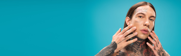 young man with tattoos touching smooth face isolated on blue, banner - Foto, Imagen