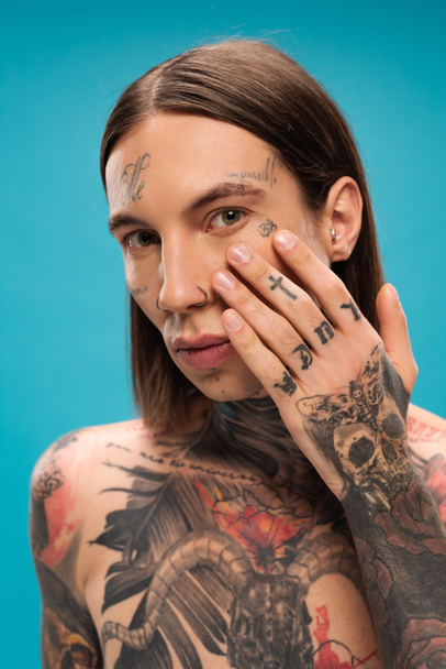 young man with tattoos applying cream under eye isolated on blue - Fotografie, Obrázek