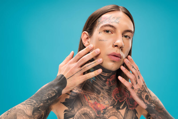 young man with tattoos touching smooth face while looking at camera isolated on blue - Foto, Bild