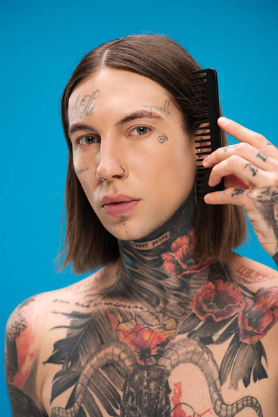 giovane e trafitto uomo con tatuaggi spazzolatura capelli con pettine isolato su blu - Foto, immagini