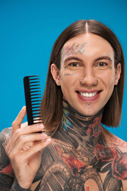 feliz e perfurado jovem com tatuagens segurando pente isolado em azul - Foto, Imagem
