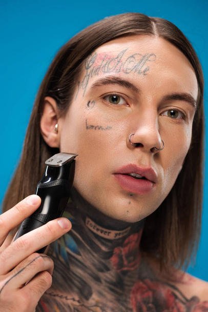 close up de homem jovem e perfurado com tatuagens de barbear com lâmina elétrica isolada em azul - Foto, Imagem