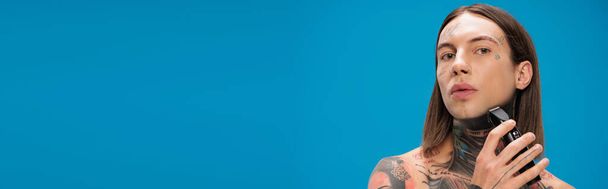 homme jeune et percé avec des tatouages rasage avec rasoir électrique isolé sur bleu, bannière - Photo, image