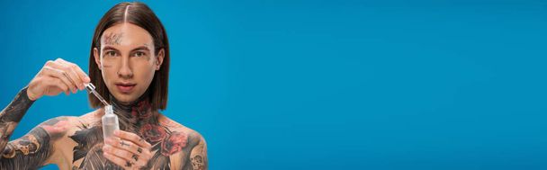 fiatal és tetovált férfi gazdaság palack hidratáló szérum izolált kék, banner - Fotó, kép