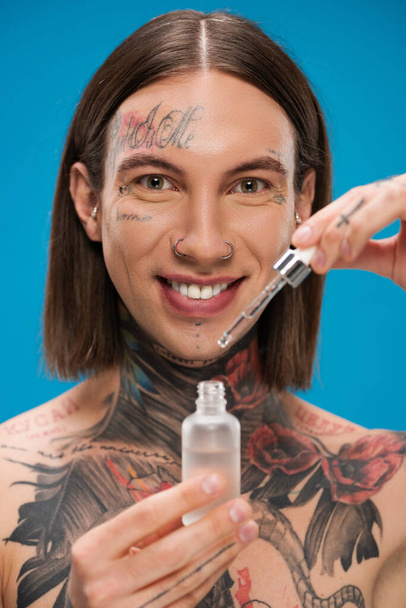 smiling and tattooed man holding bottle with moisturizing serum isolated on blue - 写真・画像