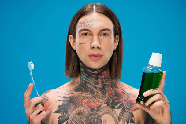 giovane e tatuato uomo in possesso di spazzolino e bottiglia con collutorio isolato su blu - Foto, immagini