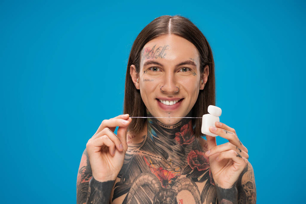 cheerful and tattooed man holding dental floss while looking at camera isolated on blue - Photo, Image