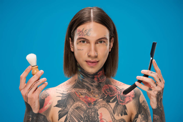 молодий татуйований чоловік тримає щітку для гоління та дзеркало ізольовано на синьому
 - Фото, зображення