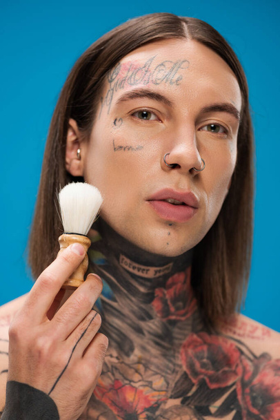 young and tattooed man holding shaving brush near face isolated on blue - Zdjęcie, obraz