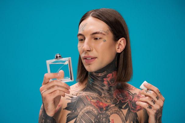 hombre joven y tatuado mirando botella con perfume aislado en azul - Foto, imagen