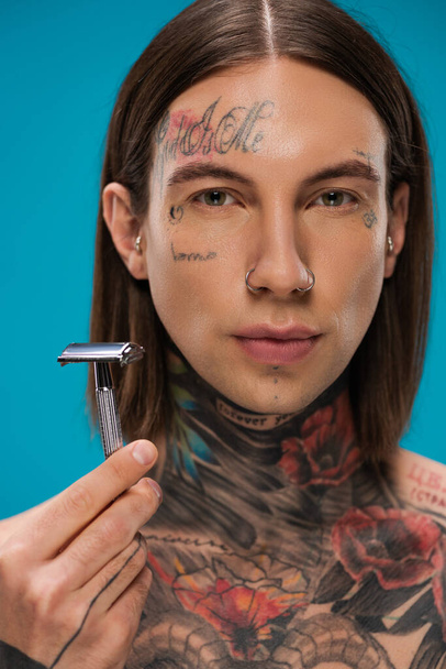 young man with tattoos holding safety razor isolated on blue - Photo, Image