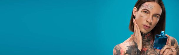 молодой человек с татуировками держа бутылку во время нанесения после бритья изолированы на синий, баннер - Фото, изображение