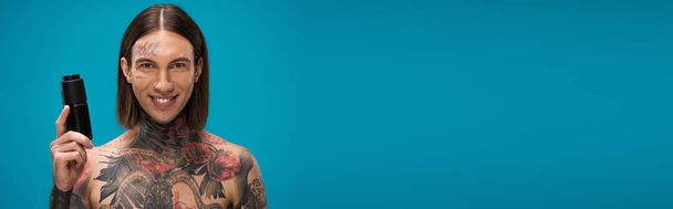 счастливый и татуированный молодой человек держит антиперспирант изолированный на голубом, баннер - Фото, изображение