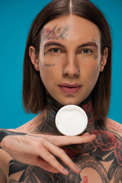 pierced young man with tattoos holding container with cosmetic cream isolated on blue  - Fotografie, Obrázek