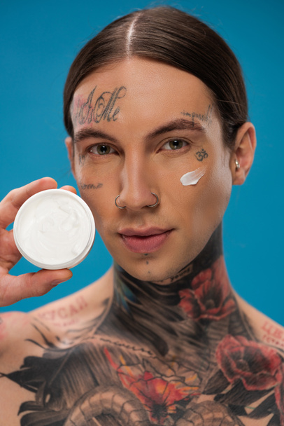 tattooed young man with cream on cheek holding container isolated on blue  - Photo, Image