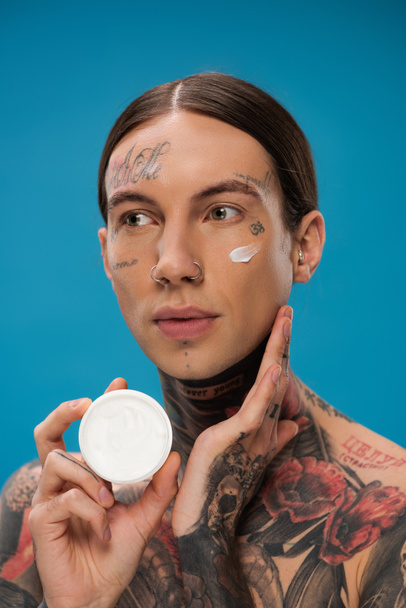 jeune homme tatoué avec crème visage sur la joue tenant récipient isolé sur bleu  - Photo, image