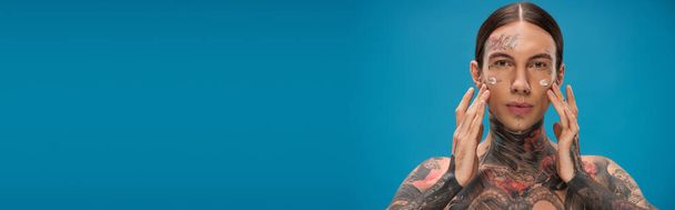tattooed young man applying cream on cheeks while looking at camera isolated on blue, banner - Foto, Imagem