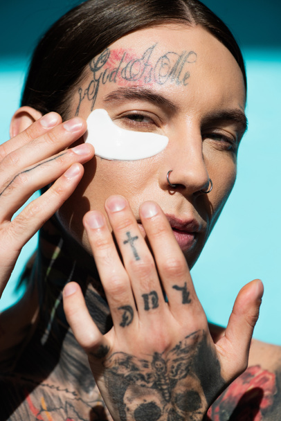 tattooed young man applying eye patch with hands and looking at camera on turquoise - Fotografie, Obrázek