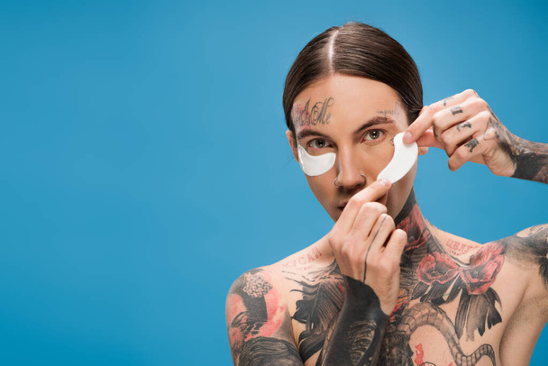 young man with tattoos applying eye patches and looking at camera isolated on blue - Photo, Image