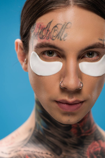 close up de jovem tatuado com manchas nos olhos olhando para a câmera isolada em azul - Foto, Imagem