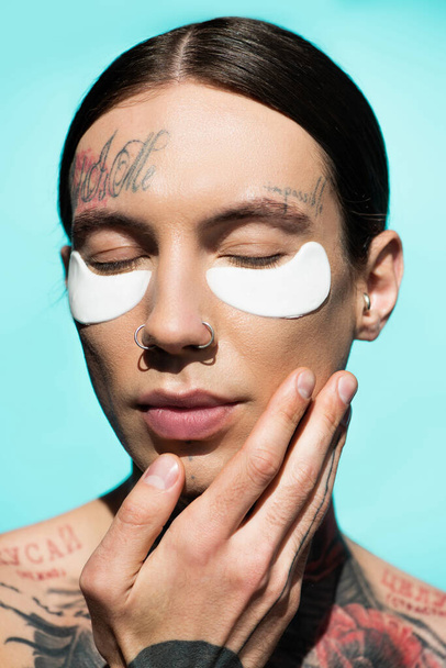 close up of tattooed young man with closed eyes and eye patches isolated on turquoise - Foto, Imagem