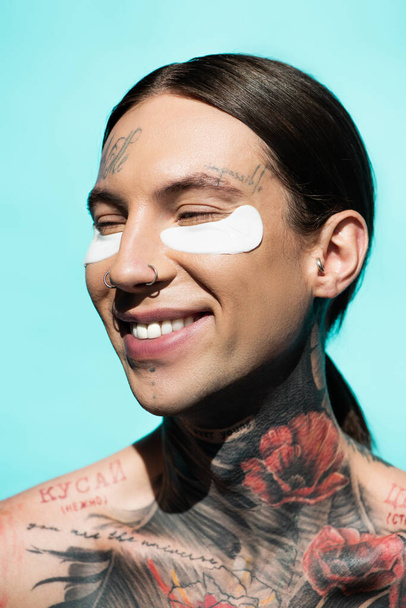 heureux et tatoué jeune homme avec des bandeaux souriant isolé sur turquoise - Photo, image