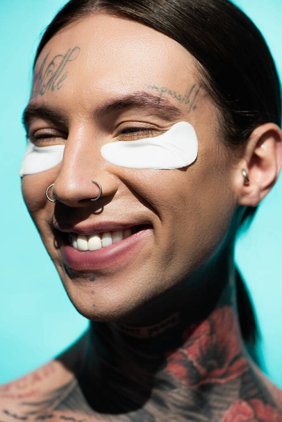 close up of tattooed young man with eye patches smiling isolated on turquoise - Photo, Image