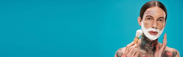 młody człowiek z tatuażami i pianką do golenia na twarzy z zabytkową szczotką odizolowaną na niebiesko, sztandar - Zdjęcie, obraz