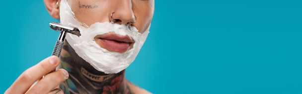 vue recadrée du jeune homme tatoué avec mousse à raser sur le visage tenant le rasoir de sécurité isolé sur bleu, bannière - Photo, image