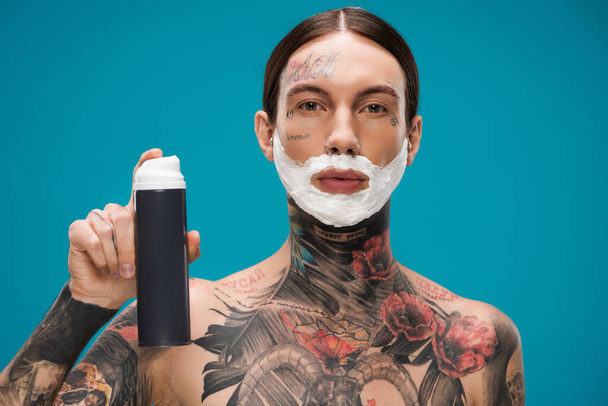 без сорочки і татуйований молодий чоловік з піною для гоління на обличчі тримає може ізольовано на синьому
  - Фото, зображення