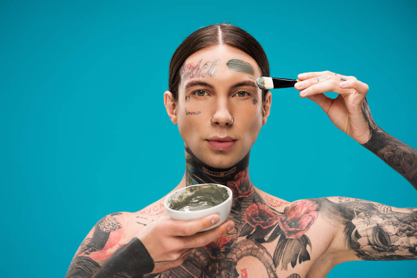 tattooed young man applying clay mask with cosmetic brush and holding bowl with product isolated on blue  - Photo, Image