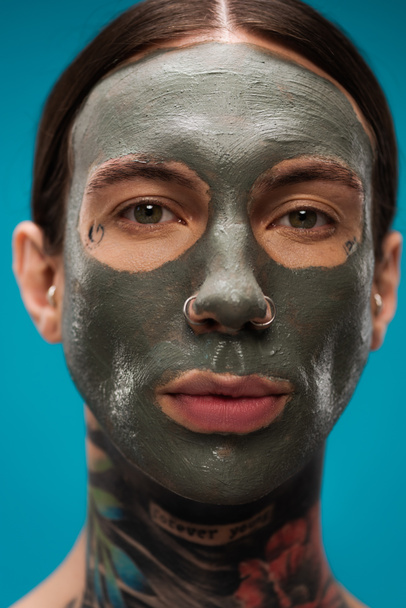 zblízka propíchnutý mladý muž s tetováním a hliněnou maskou na obličeji izolované na modré  - Fotografie, Obrázek
