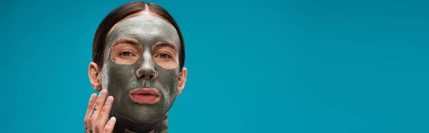 fiatal férfi tetoválásokkal megható arc agyag maszk elszigetelt kék, banner - Fotó, kép