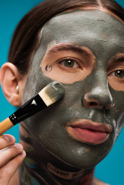 close up of pierced young man with tattoos applying clay mask with cosmetic brush isolated on blue  - Foto, Imagem