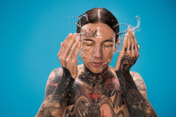 fröccsenő víz közelében fiatal tetovált férfi mosás arc elszigetelt kék - Fotó, kép