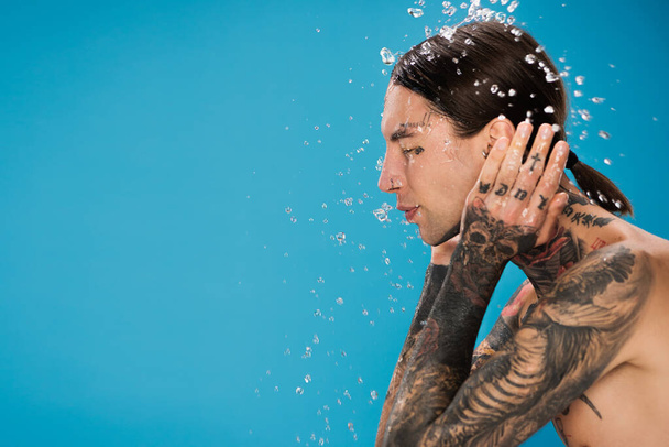 splash of water near young man with tattoos and closed eyes washing face isolated on blue - Photo, Image