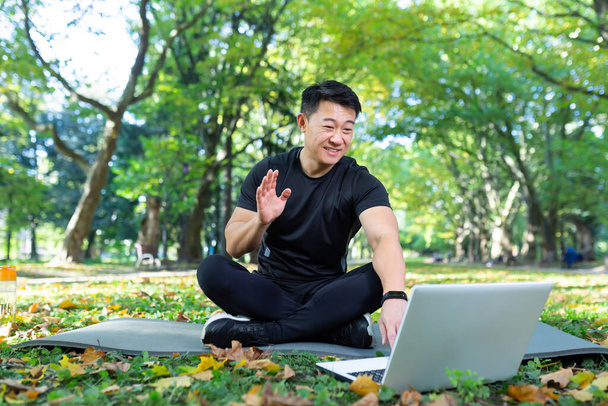 Fitness instructor works out online uses laptop in park sitting in lotus position, Asian athlete communicates with students remotely, video call training online remotely - Photo, image
