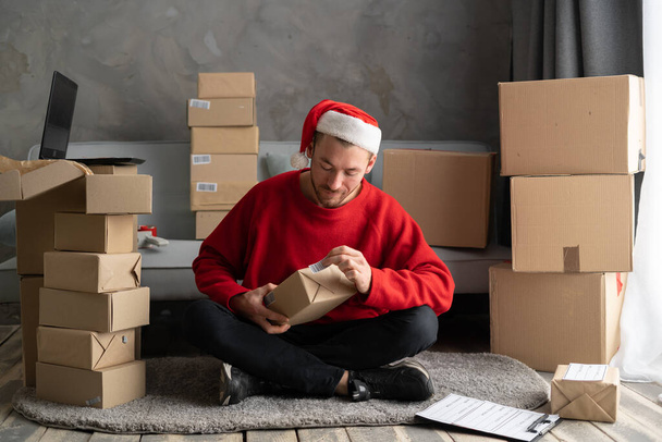 Kleinunternehmer, der im Homeoffice mit Lieferboxen auf dem Fußboden arbeitet. Mann mit Weihnachtsmütze klebt einen Strichcode auf einen Paketkasten, schnelle Lieferung zu Weihnachten. Weihnachtsverkauf und Sme-Konzept - Foto, Bild