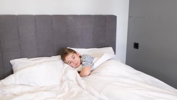 Uma linda adolescente está deitada em uma cama grande com roupa de cama branca. Bom dia. Um quarto com um interior moderno - Filmagem, Vídeo
