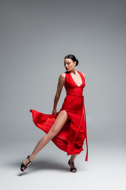 Full length of brunette dancer in red dress looking at leg on grey background - Фото, зображення
