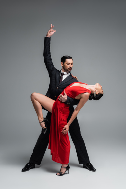 Κομψός άνθρωπος που ποζάρει ενώ εκτελεί χορογραφία tango με συνεργάτη σε γκρι φόντο - Φωτογραφία, εικόνα