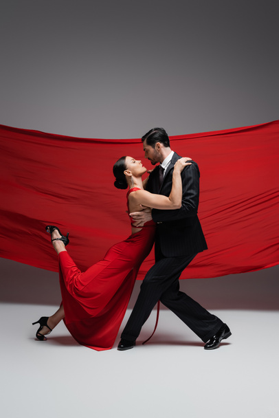 Vue latérale des danseurs de salon exécutant le tango sur fond gris avec tissu rouge  - Photo, image