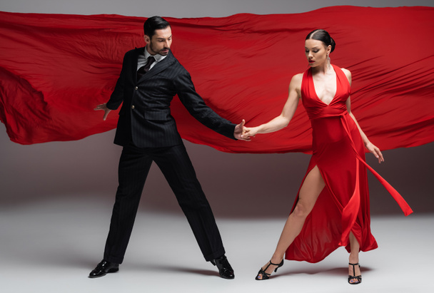 Bailarines de salón profesionales tomados de la mano sobre fondo gris con tela roja  - Foto, Imagen