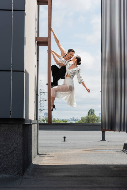 Professional ballroom dancers looking at each other while posing on ladder on rooftop of building outdoors   - Foto, immagini