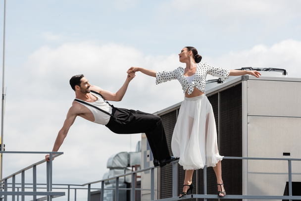 Πλευρική άποψη του χαρούμενα επαγγελματίες χορευτές κρατώντας τα χέρια κοντά κιγκλίδωμα στην ταράτσα του κτιρίου σε εξωτερικούς χώρους  - Φωτογραφία, εικόνα