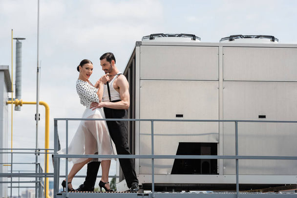 Elegante bailarina profesional en tacones mirando a la cámara mientras se mueve con su pareja en el techo  - Foto, Imagen