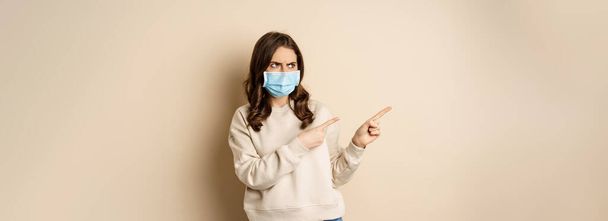 Health and covid-19 concept. Confused young woman in medical face mask, pointing left and staring with frustrated face, beige background. - Foto, afbeelding