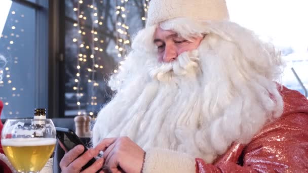 Мужчины в костюмах Санта Клауса используют смартфоны, они пьют вкусное пиво в пабе. Реклама светлого ремесленного пива. Счастливого Рождества. 4k видео - Кадры, видео